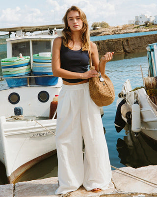 Lifestyle front view of a model wearing the LSPACE Ash Tank in midnight blue, a cropped ribbed racerback tank, styled with white wide-leg pants and a woven tote bag at a harbor setting.
